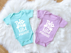 two twin baby girl or boy cotton bodysuits in lavender and mint. The bodysuits are short sleeves and they have 'two of a kind #twinlife' white print on front of both items. Bodysuits have overlap opening and are laid on a white fur background. 